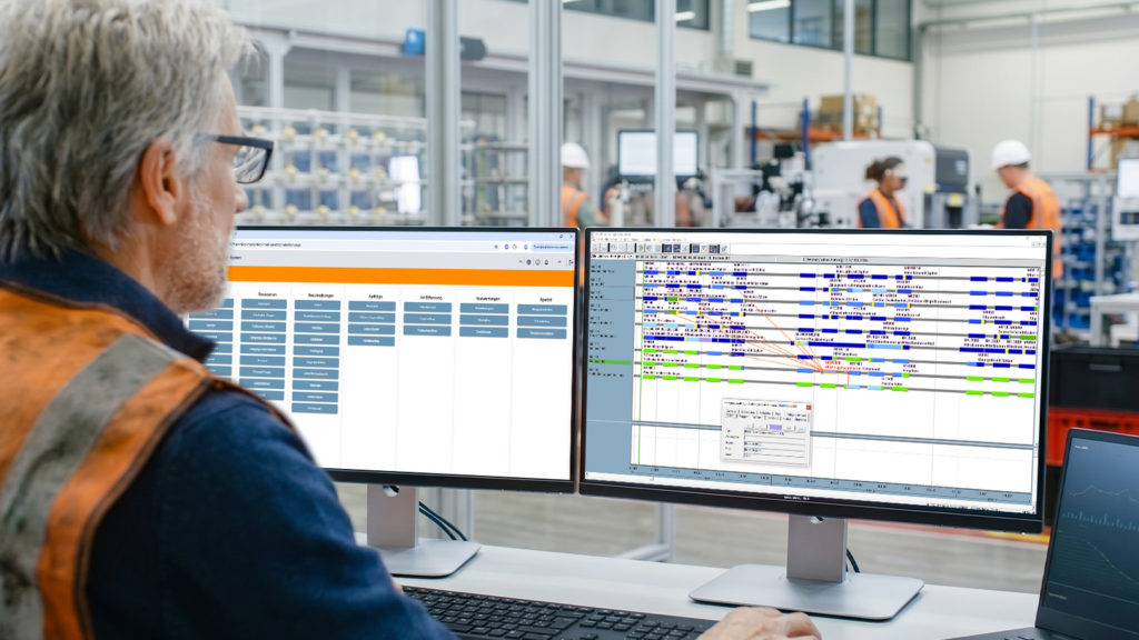 Mann mit Schutzweste vor zwei Monitoren in einer Fabrikhalle, auf den Bildschirmen Produktions- und Zeitpläne